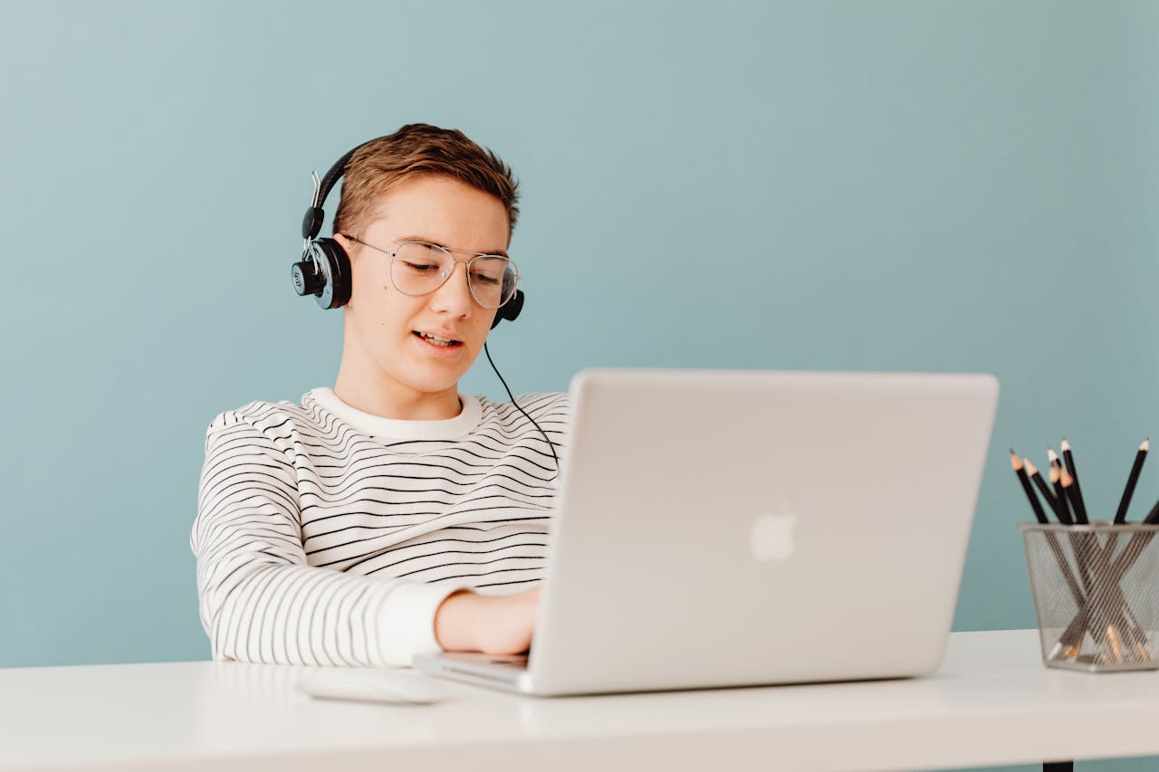 Teenager with glasses using a laptop and headphones, sitting at a table.